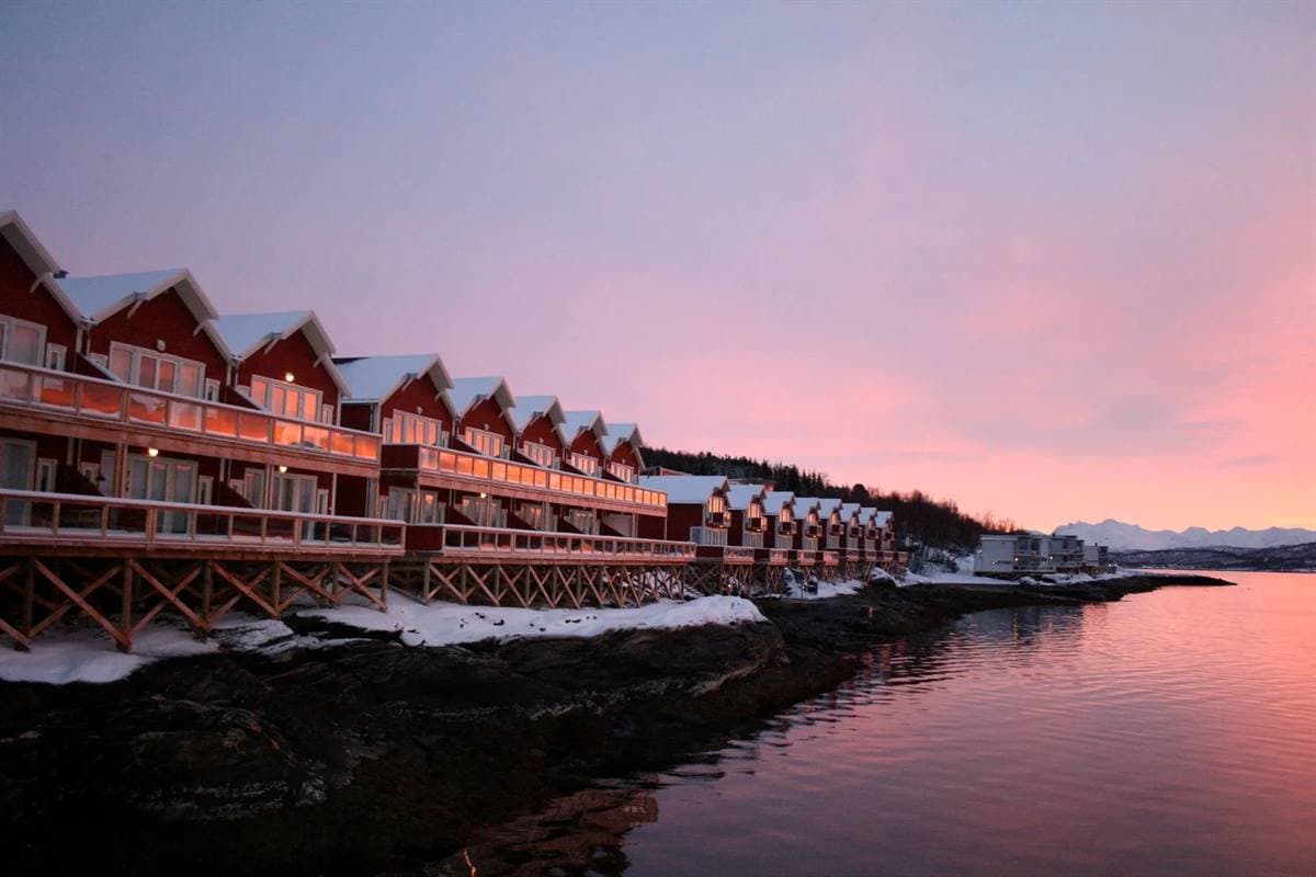 Lång röd byggnad i två våningar på pålar över den stilla fjorden i vinterskymningen