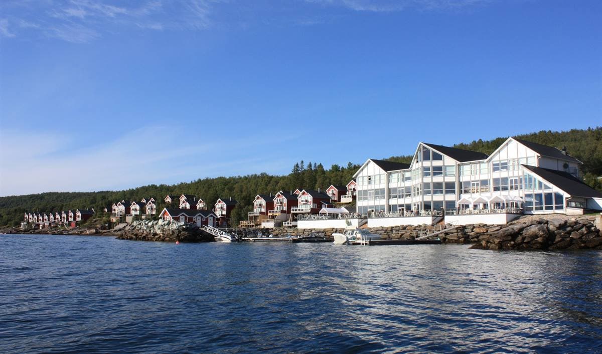 Malangen Resort sett från fjorden en sommardag, med den vita huvudbyggnaden och röda stugor längs stranden
